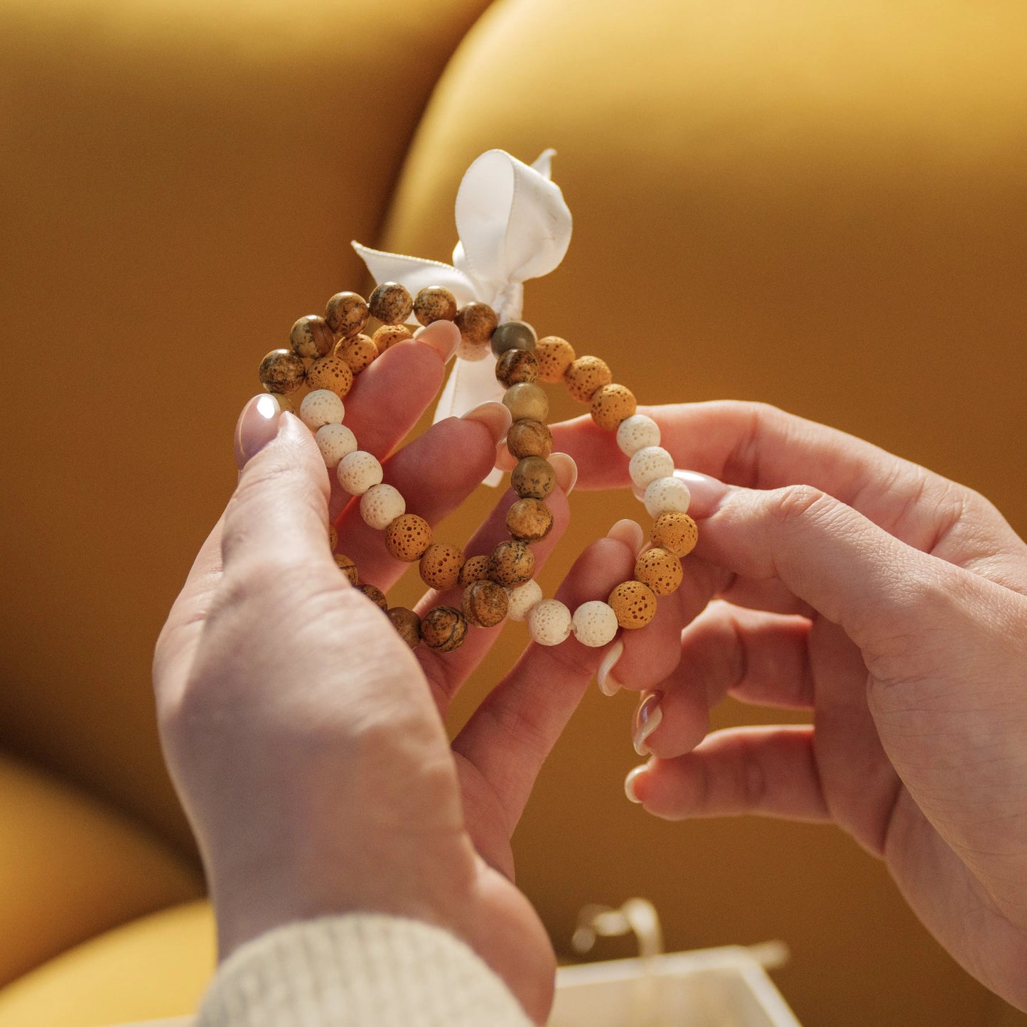 Hand holding a pair of beaded bracelets with earthy tones, tied with a white ribbon, symbolizing love and connection on a golden sofa.